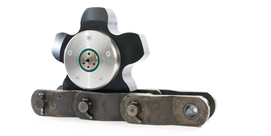 Aplicador de lubricante industrial Rotalube RA de poliacetal POM de color negro en forma de estrella, montado sobre una cadena de transmisión mecánica.